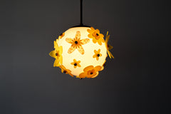 Lit floral acrylic globe pendant against a charcoal backdrop, side profile showing dimensional stacked petals in mixed yellows and chartreuse with subtle transparency, warm ambient glow and sculptural silhouette; single-edition piece designed and made in New Orleans