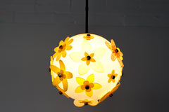Lit floral acrylic globe pendant against a charcoal backdrop, side profile showing dimensional stacked petals in mixed yellows and chartreuse with subtle transparency, warm ambient glow and sculptural silhouette; single-edition piece designed and made in New Orleans.