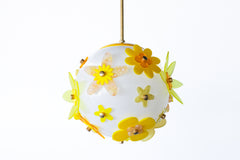 One-of-a-kind floral acrylic globe pendant light on white background, white satin-diffuse sphere wrapped in layered yellow and chartreuse acrylic flowers with warm metal caps, suspended from a warm brass stem; cheerful sculptural statement made in New Orleans.