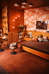 Orange Aries constellation wall sconce with four round powder-coated disc shades and glowing white globe bulbs, mounted in a warm retro bedroom with terracotta walls, oversized orange floral wallpaper, low midcentury wood platform bed, vintage wood cabinetry and shelving, and caramel striped curtains. Bold 1970s maximalist decor with a moody, eclectic vibe, perfect statement lighting for a bedroom, den, lounge, entryway, hallway, or dramatic powder room.