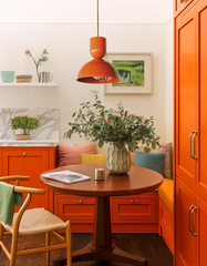 Modern breakfast nook featuring the orange Taeuber pendant light hanging above a round wood pedestal dining table with a natural woven chair and colorful banquette seating. The bright corner space includes bold orange cabinetry, marble backsplash, soft throw pillows, and greenery centerpiece, combining playful color with mid-century modern handmade lighting.