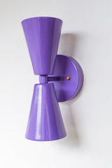 Angled view of the handmade Dryades wall sconce in orchid purple powder-coated steel, highlighting its flared dual-cone shades and brass button screw detail. This eye-catching fixture offers a modern take on vintage lighting, ideal for bathrooms, hallways, or bedrooms needing a colorful, retro-inspired accent light. Finished in a rich orchid hue for a high-impact, design-forward look.