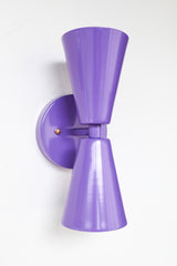 Close-up of the Orchid Dryades Sconce in a bold orchid purple finish, featuring a double-cone silhouette with a modern midcentury shape. Powder-coated steel construction with brass accents and a playful, sculptural profile. Shown mounted on a white wall, this handcrafted light adds a vibrant pop of color and sleek design to contemporary, eclectic, and maximalist interiors.