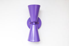 Centered view of the Orchid Dryades wall sconce with a vivid purple finish, set against a white wall for contrast. This modern handmade fixture features dual cone-shaped shades and subtle brass hardware, blending retro charm with playful contemporary design. Perfect for statement walls, kid’s rooms, or bold design lovers seeking a splash of personality in their lighting.