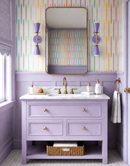 Pair of lavender Janus wall sconces with brass hardware installed beside a brass mirror above a lavender vanity with a marble countertop, colorful striped wallpaper, hex tile flooring, and brass bathroom fixtures.