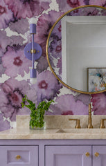 Orchid Medium Thalia Sconce shown beside a round brass mirror in a purple floral bathroom with lavender vanity cabinets, brass faucet, cream stone countertop, and oversized watercolor-style flower wallpaper. The slim vertical sconce adds colorful bathroom vanity lighting to a feminine, maximalist, grandmillennial powder room or girls bathroom design.