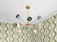 Green ikat wallpaper, white crown molding and a chandelier that does something unexpected overhead. The Orion Chandelier’s brass arms reach out in every direction, each ending in a round, open-faced shade in slate grey, vista green or cream. The shades tilt at different angles so the bulbs show, and the green ones pick up the wallpaper. A great idea for a dining room, bedroom or entry that wants pattern on the walls and color on the ceiling.