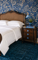 Moody blue bedroom with blue-and-white floral wallpaper, a dark wood cane headboard, crisp white bedding, and a vintage-style nightstand updated with oversized blue gumball knobs. The chunky round drawer pulls add a playful pop of color while still working with the traditional wood furniture and grandmillennial bedroom style. This image is useful for colorful furniture hardware, oversized drawer knobs, blue bedroom decor, cottage bedroom ideas, vintage nightstand makeover, and playful traditional interiors.