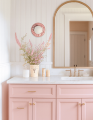 Bright airy bathroom vanity with white vertical beadboard walls and an arched natural wood mirror above a white countertop and blush pink millennial-pink cabinetry with brass hardware. A Pastel Pink Lierre ceramic wall sconce by Sazerac Stitches with a glossy pink botanical leaf relief backplate and white frosted globe light hangs above a vase of soft pink florals, paired with warm brass faucet fixtures for a modern cottage, Scandinavian coastal, and soft feminine powder room lighting look.