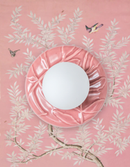 Close-up styling photo of the Pastel Pink Lierre ceramic wall sconce by Sazerac Stitches, featuring a glossy blush pink botanical leaf relief backplate and a white frosted globe light. The fixture is photographed against pink chinoiserie wallpaper with delicate white flowering branches and small birds, creating a romantic grandmillennial, chinoiserie floral, and pastel maximalist decor look for a powder room, bedroom, nursery, or feminine entryway lighting.