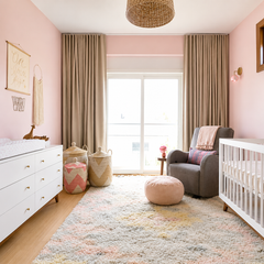 Soft blush pink nursery with a handmade Loa wall sconce in blush pink and brass, styled as a modern boho baby room. A white crib with warm wood legs sits beside a gray upholstered glider, woven ceiling pendant, and floor-to-ceiling taupe curtains. Textured pastel area rug, woven baskets, and light oak flooring create a calm Scandinavian-meets-mid-century look with warm, feminine color and brass accents.