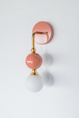 The Ida Sconce in peach stacks a hooked brass arm, ribbed brass ball joints, and a glossy peach powdercoated sphere above a soft white opal glass globe, giving the whole fixture a jewelry-like silhouette on the wall. It works well as a bedside light in place of a table lamp, beside a bathroom mirror, or anywhere a single statement sconce makes more sense than a matching pair.
