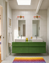 Bright modern bathroom with two peach and brass Janus sconces mounted above twin mirrors, adding playful geometric lighting to a clean white tiled wall. A floating green double vanity, white vessel sinks, chrome faucets, penny tile floor, skylight, and rainbow bath rug make the space feel fresh, colorful, and family-friendly. The peach wall sconces bring a soft dopamine decor moment to this kids bathroom or modern powder room while still working with crisp, minimalist tile and simple architectural lines.