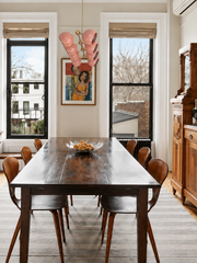 This city dining room earns its mix: a dark farmhouse-leaning table with molded plywood chairs, a carved antique hutch, figurative art, and tall black-framed windows looking over the rooftops. Overhead, the Calliope chandelier in Peach and Brass clusters glossy peach globes on a geometric brass armature, the one playful, sculptural gesture in a room of serious wood. Statement dining room lighting for collected, eclectic homes that mix eras on purpose.