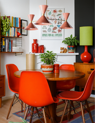 Alt Text (SEO-heavy)
Playful mid-century dining room featuring the Peach & Brass Clancy chandelier with cone-shaped shades, hanging above a round wood dining table surrounded by red Eames-style chairs. The retro-inspired space includes colorful bookshelves, bold red ceramic accents, houseplants, a green table lamp, and graphic wall art, blending vintage charm with modern handmade lighting design.