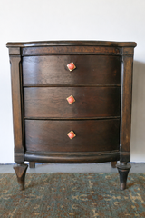 A dark wood nightstand with a bowed drawer front and turned, tapered legs gets an easy update with peach and brass gumball knobs on each drawer. The glossy peach pulls read fresh and juicy against the deep, aged wood tone, proof that a small hardware swap can bring real personality to an older piece without refinishing it. Sitting on a faded vintage-style rug, the whole vignette leans collected rather than matched.