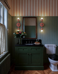 Lit peach and chrome Medium Thalia Sconces shown as a pair of vertical vanity lights flanking a beaded wood mirror in a moody olive green bathroom. The space features green beadboard walls, a matching painted vanity, black stone countertop, pink and tan striped wallpaper, café curtains, wood herringbone floors, and old-house details. A colorful bathroom sconce idea for vintage powder rooms, traditional bathrooms, cottage interiors, playful striped wallpaper, olive green vanities, and warm vanity lighting wi