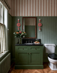 Peach and chrome Medium Thalia Sconces installed on either side of a bathroom mirror in an olive green powder room with classic beadboard paneling, a green vanity, black countertop, striped wallpaper, antique-style wood floors, and a vintage beaded mirror. The slim vertical sconces add a playful pop of peach against the traditional green walls, making them a great choice for colorful bathroom lighting, small powder rooms, grandmillennial bathrooms, old-house renovations, striped wallpaper bathrooms, and ecl
