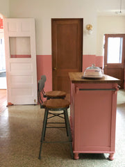 A kitchen with a bold, personalized aesthetic featuring dusty rose wainscoting on the lower half of walls and cream above. A custom pink painted island with turned wooden feet and natural wood countertop serves as the centerpiece, flanked by two wooden stools with dark metal frames. Brass articulating West End sconces mounted on the cream wall provide focused task lighting. Brown wood cabinetry and doors, along with white painted doors, define the space. A glass dome cloche and natural wood accessories sit 