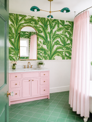 This bathroom goes all in on a pink and green tropical palette, wrapping the upper walls in a classic banana leaf print and painting the vanity a candy pink to match. The Coquille Chandelier overhead picks up the same mermaid green in its leaf-shaped shades, with brass arms and fixtures warming up the whole room. A green paneled door, an arched brass mirror, and a striped shower curtain round out a bathroom that feels collected and playful rather than themed.
