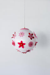 Product photo of the Pink and Red Blossom Gumball Chandelier on a clean white background, a 16 inch white globe pendant decorated with layered acrylic flowers in blush pink, hot pink, and red with brass center caps, a whimsical modern pendant light for maximalist homes, nurseries, and playful interiors.