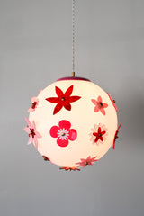 Close up of the Pink and Red Blossom Gumball Chandelier while illuminated, showing the warm glow through the white globe shade and the layered acrylic flower appliqués in pink and red with brass center hardware, a bold statement pendant light for dopamine decor, retro inspired interiors, and creative kids spaces.