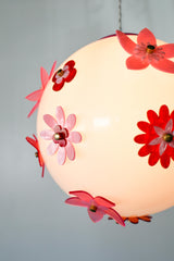 Side angle lit photo of the Pink and Red Blossom Gumball Chandelier featuring a glowing white globe pendant with scattered acrylic flower appliqués in hot pink, translucent pink, and deep red with brass centers, a fun modern flower power ceiling light for eclectic dining rooms, playful bedrooms, and maximalist interiors.