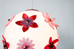 Side angle lit photo of the Pink and Red Blossom Gumball Chandelier featuring a glowing white globe pendant with scattered acrylic flower appliqués in hot pink, translucent pink, and deep red with brass centers, a fun modern flower power ceiling light for eclectic dining rooms, playful bedrooms, and maximalist interiors.