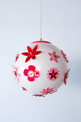 Full view of the Pink and Red Blossom Gumball Chandelier illuminated, showing a warm glowing white globe shade covered in dimensional acrylic flower appliqués in pink and red with brass center details, a bold flower power statement pendant light for eclectic interiors, colorful dining rooms, and kids spaces.