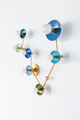 Studio product photo of Pisces zodiac sconce with constellation layout, powder-coated steel discs in mixed blues and greens paired with warm brass hardware and white globe bulbs; bold celestial wall lighting for a statement powder room, colorful bathroom vanity lighting, funky modern entryway sconce, or playful kids bedroom decor, perfect for design lovers who want artful, sculptural, mid century modern inspired lighting.