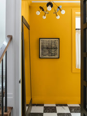 A color-drenched mustard yellow hallway meets a dramatic black-and-white checkerboard floor, black doors, and graphic architectural artwork for a bold modern entryway. Overhead, the customizable Pitot Chandelier by Sazerac Stitches features six exposed globe bulbs, slender brass arms, and circular disc shades in black, brass, and cool metallic finishes. The sculptural ceiling light adds playful geometry to this high-contrast interior—a distinctive idea for colorful hallway lighting, postmodern decor, and st