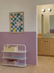 Playful modern bathroom with Vista Green Abeille sconces above a clean mirror and pale green vessel sink, set against a soft pastel purple and white color-blocked wall. The plywood vanity, plywood flooring, simple storage cart, checkerboard flower art, and pastel crates give the space a kid-friendly, Scandinavian, minimalist, and dopamine decor feel. A sweet example of colorful bathroom lighting for a kids bathroom, family bath, modern powder room, or design-forward pastel interior.