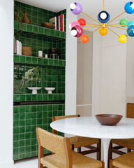 Rainbow Prism Chandelier featuring 12 vibrant powder-coated globes on a brass sputnik frame, styled in a mid-century modern dining room with a white tulip table, woven wood chairs, and dramatic green tiled shelving — a bold eclectic lighting choice for color lovers.