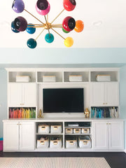 Prism Chandelier in a modern playroom with custom white built-ins, rainbow color-coded books, woven storage baskets, and a mounted TV. Mid-century modern rainbow sputnik ceiling light adds dopamine décor style and playful eclectic energy to kids’ spaces and creative family rooms.
