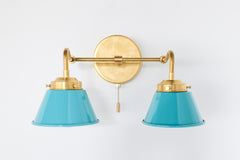 Double Kelly wall sconce in brass with teal shades and a pull chain switch, mounted on a white wall.