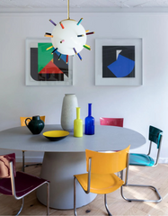 Playful modern dining room featuring the Rainbow Spike Gumball pendant light, a sculptural globe fixture with multicolor spikes, hanging above a round gray pedestal dining table. The eclectic space includes bold colored chairs, minimalist artwork, and vibrant glass vases, blending contemporary statement lighting with artistic, retro-inspired interior design.