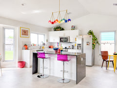 Wide shot of a bright, open white kitchen with a rainbow Turmalina chandelier as the main statement piece over the island. The colorful linear chandelier adds playful maximalist energy to the white cabinetry, gray island, pink acrylic stools, yellow breakfast table, plants, and cheerful accessories throughout the room. This image works for modern kitchen lighting, colorful chandeliers, rainbow decor, eclectic kitchen design, dopamine decor, and bold lighting ideas for bright white interiors.