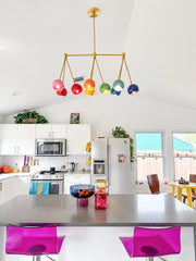 Bright white kitchen with a rainbow Turmalina chandelier hanging over a large gray island, bringing bold color to a clean modern space. The brass linear frame holds colorful glass shades in pink, red, orange, yellow, green, blue, and purple, pairing beautifully with hot pink acrylic bar stools, white cabinets, subway tile, plants, and playful countertop accessories. A strong fit for colorful kitchen lighting, dopamine decor, maximalist interiors, modern brass chandeliers, and statement lighting for a white 