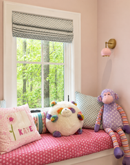 Soft blush pink reading nook featuring a built-in window seat styled with playful pillows and stuffed animals, plus a blush pink Loa wall sconce with warm brass hardware. The glossy dome shade and round brass backplate add a modern, mid-century inspired touch while casting cozy light for bedtime stories. White window trim, a patterned Roman shade, and leafy outdoor views create an airy, sweet kids space that blends cottage charm with modern nursery design. Great inspo for girls bedroom lighting, nursery wal