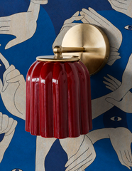Cobalt blue wallpaper covered in graphic cream hands and eyes brings a surrealist, gallery-like energy to this wall, and the Marcella Sconce in Jasper Red holds its own against it. The fluted red shade and brass ball finial read as sculptural rather than competing, proof that a bold printed wallpaper and a saturated colored sconce can share a wall. Maximalist powder room inspiration for anyone drawn to art-forward, conversation-starting wallpaper.