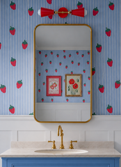 This bathroom takes a strawberry-print wallpaper and runs with it, pairing periwinkle blue stripes with a matching blue vanity and warm brass hardware. Above the gold-framed mirror, a red horizontal Janus Sconce glows with a matching pop of color, its double-cone shape giving the room a playful and flirty, cheeky, mid-century edge. Framed strawberry and floral prints add a collected, personal touch, proving a fruit-print bathroom can feel design-forward instead of juvenile.