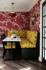 A breakfast nook that commits fully to pattern: red and cream toile wallpaper wraps the walls behind a built-in banquette upholstered in mustard floral chintz, with a marble bistro table on a black iron base pulled up close. A wine colored fluted pendant light on a brass stem hangs in the corner, warm and jewel toned against all that red. It’s chinoiserie traditional done with confidence, the kind of small, saturated space that makes a whole house feel collected.