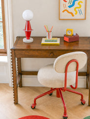 A playful eclectic study space featuring the red Totem Lamp styled on a dark wood writing desk with traditional lines and carved edge detailing. The room pairs classic furniture with colorful, postmodern accents, including a cream upholstered rolling desk chair with a red metal frame, bright desk accessories, and whimsical wall art. The Totem Lamp adds sculptural personality with its glossy red geometric base, white globe shade, and marble foot, making it a standout accent for a kids desk, homework station,