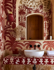 This powder room leans into old-world Mediterranean charm: red and cream palm print wallpaper climbs the walls and wraps the vanity ledge, framing an arched wood door and a sculptural white vessel sink with black wall-mount fixtures. The Marcella Sconce in Jasper Red picks up the wallpaper's deepest red, its fluted shade and brass ball finial adding warmth beside the mirror. Tropical traditional powder room inspiration for anyone who wants pattern with real texture and history.