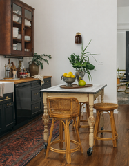 This kitchen earns its lived-in feel honestly: black shaker cabinets with brass hardware, a chippy painted table with a marble top, mismatched rattan counter stools on casters, and an antique-style runner worn in just right. The Marcella Sconce in Chicory Brown glows warmly against the plain white wall, its fluted glossy shade fitting right into all that collected patina. Farmhouse kitchen inspiration for anyone who wants character over polish.