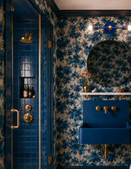 Blue and brass Medium Thalia Sconce installed horizontally over a round bathroom mirror in a moody floral wallpaper powder room. The saturated blue vanity light coordinates with the blue wall-mounted sink, blue tile shower, navy trim, and botanical wallpaper, while the brass accents tie into the shower hardware, faucet, and shelf brackets. A playful modern wall sconce for maximalist bathrooms, small powder rooms, colorful kids bathrooms, blue-and-gold decor, and statement vanity lighting.