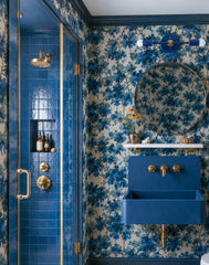 Medium Thalia Sconce in Mediterranean Blue and brass shown as a horizontal bathroom vanity light above a round mirror in a dramatic blue floral bathroom. The room features sapphire blue botanical wallpaper, dark blue trim, glossy blue shower tile, brass plumbing fixtures, a wall-mounted blue sink, and layered maximalist style. A colorful two-light wall sconce for bold powder rooms, blue bathrooms, grandmillennial interiors, floral wallpaper bathrooms, eclectic vanity lighting, and brass bathroom fixture pai