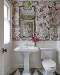 This powder room wraps the walls in a black-and-white rose print scattered with colorful printed butterflies, serious and playful at the same time. An ornate curved black mirror anchors the pedestal sink below, while the Sid Sconce mounted above it brings one confident pop of chartreuse and gold to break up all that black and white. A diamond-pattern marble floor and a vase of fresh magenta flowers keep the room feeling finished rather than themed.
