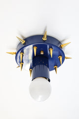 Product photo of the Sid Sconce in deep navy with gold-tone spikes, shown mounted as a compact wall light. The angled composition emphasizes the hand-drilled spike placement, glossy powder-coated finish, white globe bulb, and the fixture’s playful but slightly rebellious design.