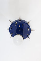 Side-angle product photo of the Sid Sconce in glossy navy with silver-tone spikes, mounted as a small wall sconce against a simple white background. The image shows the fixture’s dimensional shape, projecting bulb, spiked round backplate, and bold mix of playful color with sharper metal accents.